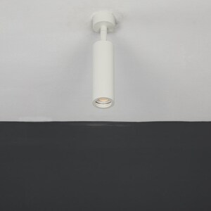 May include: A white cylindrical ceiling light fixture with a single light bulb. The light is mounted on a white ceiling and is pointing downwards.