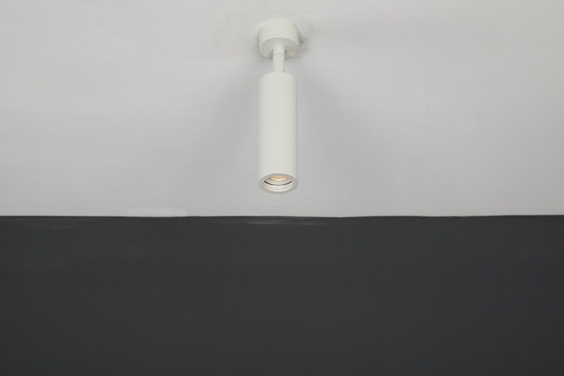 May include: A white cylindrical ceiling light fixture with a single light bulb. The light is mounted on a white ceiling and is pointing downwards.
