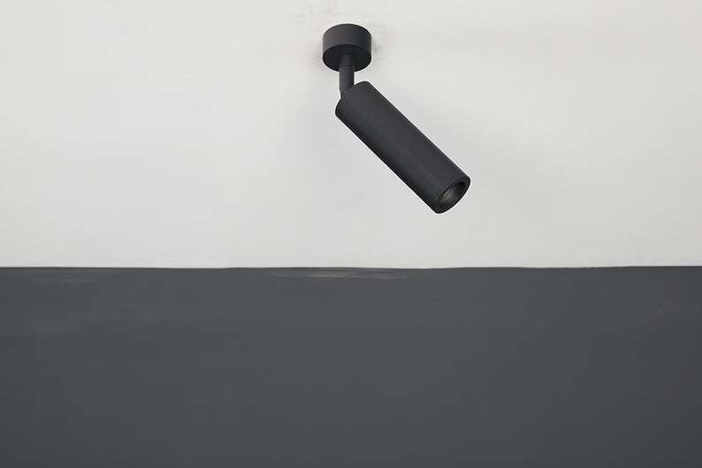 May include: A black ceiling light fixture with a cylindrical shade angled downwards. The light is mounted on a white ceiling.