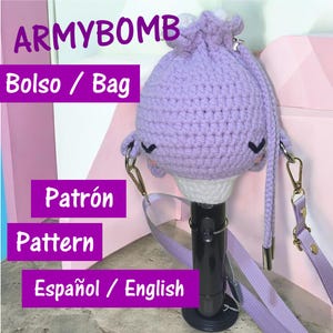 May include: A purple crocheted bag with a cartoon face design, a strap, and metal clasps. The bag is designed to hold a black object. Text on the image reads "ARMYBOMB", "Bolso / Bag", "Patrón / Pattern", and "Español / English".