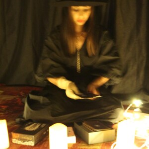 May include: A dark scene with a person in a black hat and dress, sitting on a patterned rug. Candles and fairy lights illuminate the area, with a box and cards present. The image evokes a mystical or spiritual atmosphere.