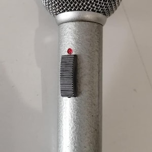 May include: A silver-colored microphone with a mesh head and a cylindrical body. It features a black slider switch and a red indicator light. The microphone has the brand name "Beyer" printed on it.