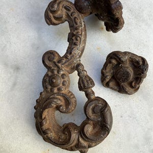 Peut inclure: Gros plan sur trois pièces de quincaillerie anciennes en fonte ornée. Les pièces sont brun foncé avec une patine rouillée. L'une est une poignée décorative, une autre est un bouton et la troisième est un élément décoratif.