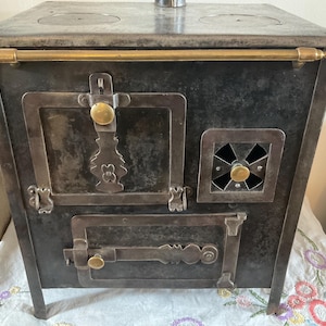May include: An antique metal stove with a dark finish, featuring brass accents on the handle and knobs. The stove has two circular burners on top, a small vent, and two doors with ornate latches. It stands on four legs.