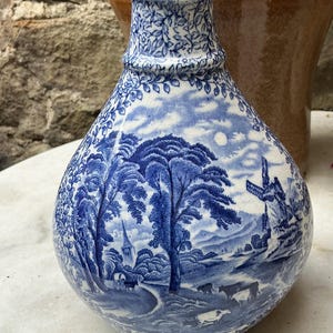 May include: Blue and white ceramic vase with a landscape scene. The vase features a detailed illustration of trees, a church, and a windmill. The top of the vase has a floral pattern. The vase is a decorative item.