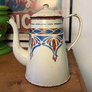 Op de afbeelding: Oude witte emaille koffiepot met een decoratief blauw en bruin bloemmotief. De pot heeft een gebogen tuit, een handvat en een deksel. De rand en het deksel zijn rood afgewerkt. Een charmant keukenartikel.