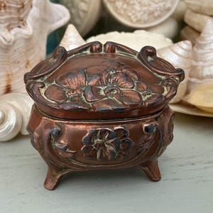 May include: A small, antique copper-coloured jewellery box with floral relief detailing. The box has a hinged lid and rests on three small feet. The lid and body feature embossed flower designs. The box is likely made of metal and has an aged patina.