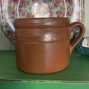 May include: A cylindrical brown stoneware mug with a thick, rounded rim and a single sturdy handle on the right side. The surface of the mug has a smooth, matte finish with subtle variations in the clay color, featuring a horizontal ridge.