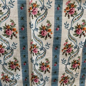 Op de afbeelding: Close-up van een stof met een herhalend bloemenpatroon. Het ontwerp toont roze en oranje rozen met groene bladeren en blauwe linten tegen een witte achtergrond met blauwe verticale strepen. De stof heeft een getextureerde uitstraling.