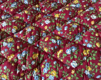 Puede incluir: Primer plano de una tela acolchada con un estampado floral. La tela es de color burdeos intenso, con un patrón repetido de flores azules y amarillas y hojas verdes. El acolchado crea un patrón de diamantes.