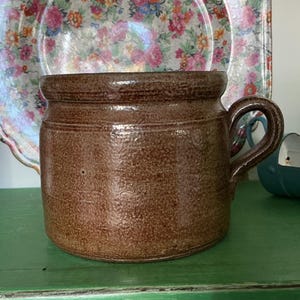 Può includere: Una tazza in ceramica marrone con una finitura testurizzata e maculata, dotata di un manico spesso e arrotondato. Il corpo cilindrico presenta un bordo leggermente svasato e dettagli a scanalature orizzontali vicino alla parte superiore.