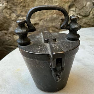 May include: A dark, weathered metal weight with a conical body and a flat top. It features a central arched handle flanked by two decorative, spool-shaped finials. A hinged locking mechanism extends across the top, securing the lid to the base.