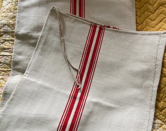 May include: Two white linen tea towels with red stripes. The towels are folded and stacked on top of each other.