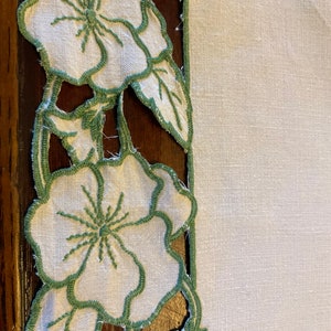 Puede incluir: Mantel de mesa de lino blanco con un borde floral bordado en verde. Las flores son estilizadas y tienen un aspecto vintage.