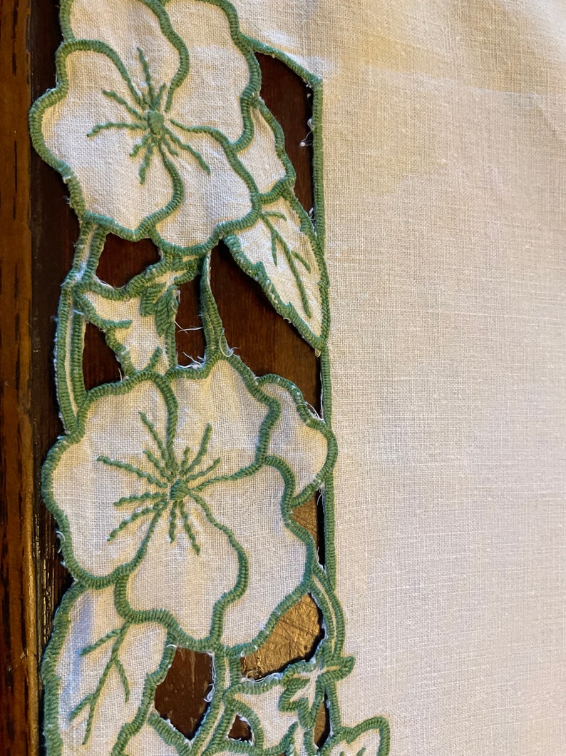 Puede incluir: Mantel de mesa de lino blanco con un borde floral bordado en verde. Las flores son estilizadas y tienen un aspecto vintage.