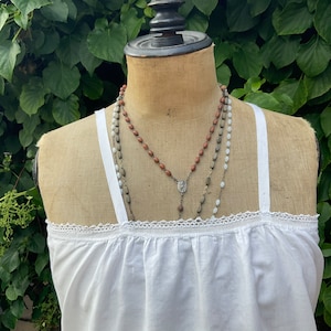 Può includere: Un top bianco in cotone senza maniche con un bordo in pizzo e tre collane sovrapposte. Le collane sono fatte di perline bianche, marroni e argentate.