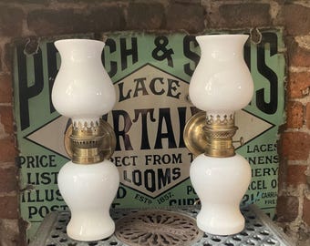 Op de afbeelding: Twee antieke witte glazen wandlampen met messing accenten. De lampen hebben een klassiek ontwerp met een afgeronde basis en een uitlopende bovenkant. Een vintage bord staat op de achtergrond.