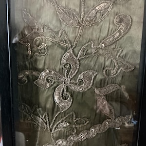 Könnte beinhalten: Gerahmtes Kunstwerk mit olivgrünem Stoff und aufwendiger silberner Stickerei. Die Stickerei zeigt Blumen- und Blättermuster, verziert mit Pailletten. Das Kunstwerk ist in einem schwarzen Rahmen eingefasst, was seine visuelle Wirkung verstärkt.