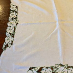 Puede incluir: Mantel de mesa de lino blanco con un borde floral bordado en verde. Las flores est&aacute;n recortadas y el dise&ntilde;o es intrincado.