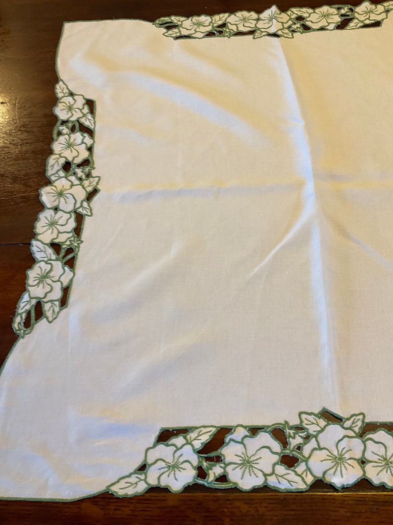 Puede incluir: Mantel de mesa de lino blanco con un borde floral bordado en verde. Las flores est&aacute;n recortadas y el dise&ntilde;o es intrincado.
