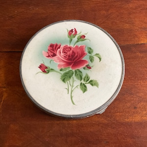 Könnte beinhalten: Runder Kompaktspiegel mit floralem Design. Der Kompaktspiegel hat einen silberfarbenen Metallrand und eine cremefarbene Oberfläche mit einem Gemälde einer rosa Rose und Knospen mit grünen Blättern. Der Hintergrund ist hellblau.
