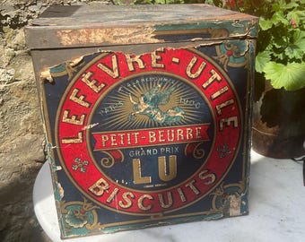 May include: Vintage biscuit tin with the text "LEFEVRE-UTILE PETIT-BEURRE BISCUITS" in red and gold lettering on a dark blue background. The tin has a rectangular shape with a hinged lid and shows signs of wear.