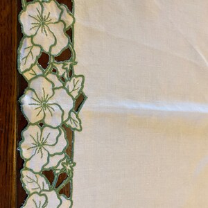 Puede incluir: Mantel de mesa de lino blanco con un borde floral bordado en verde. El borde presenta un patr&oacute;n repetitivo de flores y hojas.