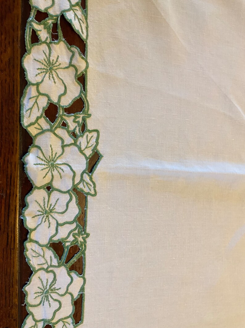 Puede incluir: Mantel de mesa de lino blanco con un borde floral bordado en verde. El borde presenta un patr&oacute;n repetitivo de flores y hojas.