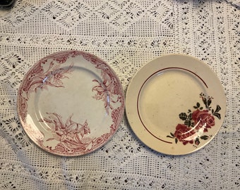 May include: Two vintage ceramic plates with floral designs. One plate features a pink floral pattern with a white background, while the other plate has a red rose design with a cream background.