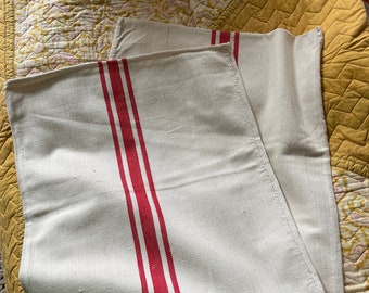 May include: Two white linen tea towels with red stripes. The towels are folded and placed on a yellow and white patterned fabric.