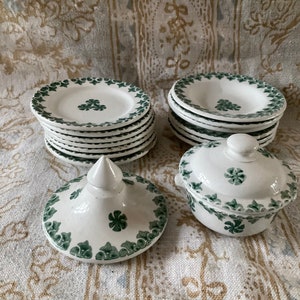 May include: A set of white ceramic plates with green clover leaf designs. The plates are stacked in two piles, and there is a small white ceramic bowl with a lid and a green clover leaf design.