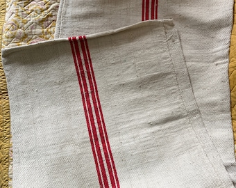 May include: Two white linen tea towels with red stripes. The towels are folded and stacked on top of each other.