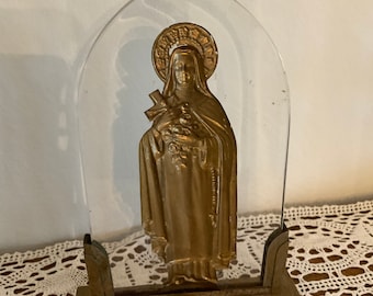 May include: A gold-colored religious figurine of a woman in a robe, with a halo and a cross, is displayed in a clear plastic frame on a gold-colored metal base.