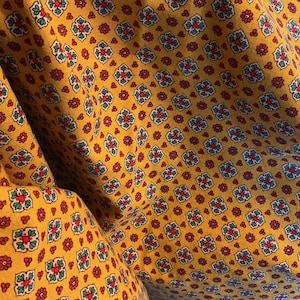 May include: Close-up of a yellow fabric with a repeating floral pattern. The design features red, green, and blue floral and geometric shapes. The fabric is folded, creating shadows and depth, highlighting the texture and pattern.