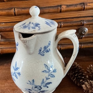 May include: A cream-colored ceramic pitcher with a matching lid, featuring a delicate blue floral pattern across the surface. The pitcher has a curved handle, a small spout, and a visible crackled glaze finish throughout the entire piece.