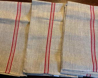 May include: Three folded linen kitchen towels. The towels are a light gray color with three vertical red stripes. The fabric has a slightly textured appearance.