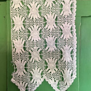 以下が含まれることがあります： 白い手作りのかぎ針編みカーテンパネル。繰り返し模様の花柄が特徴です。長方形のパネルはスカラップエッジで、緑色の背景に表示されています。花柄のデザインが主な特徴です。