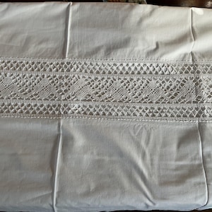 May include: White cotton fabric with a wide, decorative band of white lace. The lace features an openwork design with floral and geometric patterns. The fabric is folded and appears to be a tablecloth or bedding.