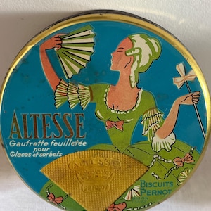 Puede incluir: Lata redonda vintage con fondo turquesa y borde dorado. La lata presenta a una mujer con un vestido verde sosteniendo un abanico. El texto "ALTESSE" se muestra en letras doradas, junto con otro texto francés y una oblea dorada.