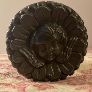 May include: A black cast iron decorative knob featuring a cherub face surrounded by a floral design.