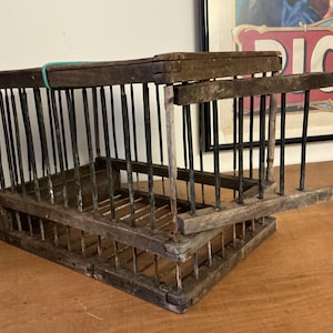 Pode incluir: Uma gaiola de pássaro de madeira antiga com barras de metal verticais. A gaiola é retangular com uma porta articulada e mostra sinais de desgaste. Uma faixa de borracha verde prende a parte superior. A gaiola está em uma superfície de madeira.