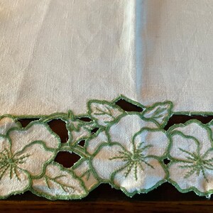 Puede incluir: Mantel de mesa de lino blanco con un borde floral bordado en verde. Las flores son estilizadas y tienen un borde festoneado.