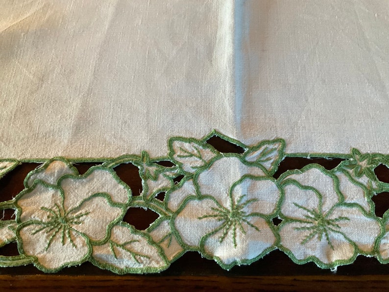 Puede incluir: Mantel de mesa de lino blanco con un borde floral bordado en verde. Las flores son estilizadas y tienen un borde festoneado.