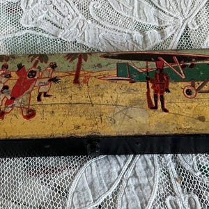 Op de afbeelding: Een rechthoekige zwarte metalen doos met een vintage illustratie op het deksel. De scène toont een groen vliegtuig in een geel woestijnlandschap met palmbomen, een figuur in een rood uniform met een geweer en een groep mensen in witte gewaden.