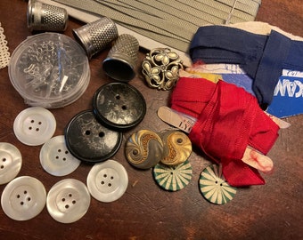 Op de afbeelding: Een verzameling naaigerei, waaronder knopen, spoelen met lint, vingerhoeden en een decoratieve metalen pin. De items zijn gerangschikt op een houten oppervlak.