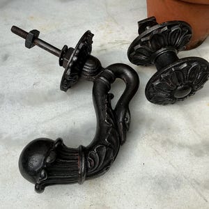 May include: Ornate black metal drawer pulls and handles. The hardware features a detailed floral design and a curved handle. One handle is attached to a bolt with a nut and washer. The other two pieces are separate.