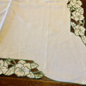 Puede incluir: Mantel de mesa de lino blanco con un borde floral bordado en verde. El bordado presenta un patr&oacute;n repetitivo de flores y hojas.