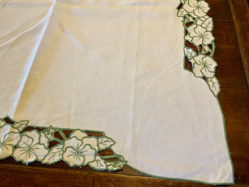 Puede incluir: Mantel de mesa de lino blanco con un borde floral bordado en verde. El bordado presenta un patr&oacute;n repetitivo de flores y hojas.