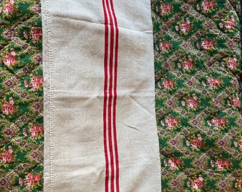 May include: A white linen tea towel with three red stripes. The towel is folded in half and lying on a green floral patterned surface.