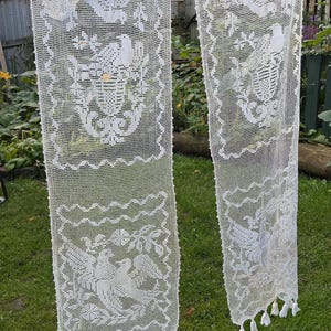 May include: Two long, rectangular white lace panels featuring intricate crochet patterns of birds and floral motifs. Each panel has a grid-like texture, decorative borders, and small tassels hanging from the bottom edge, suspended by clothespins.